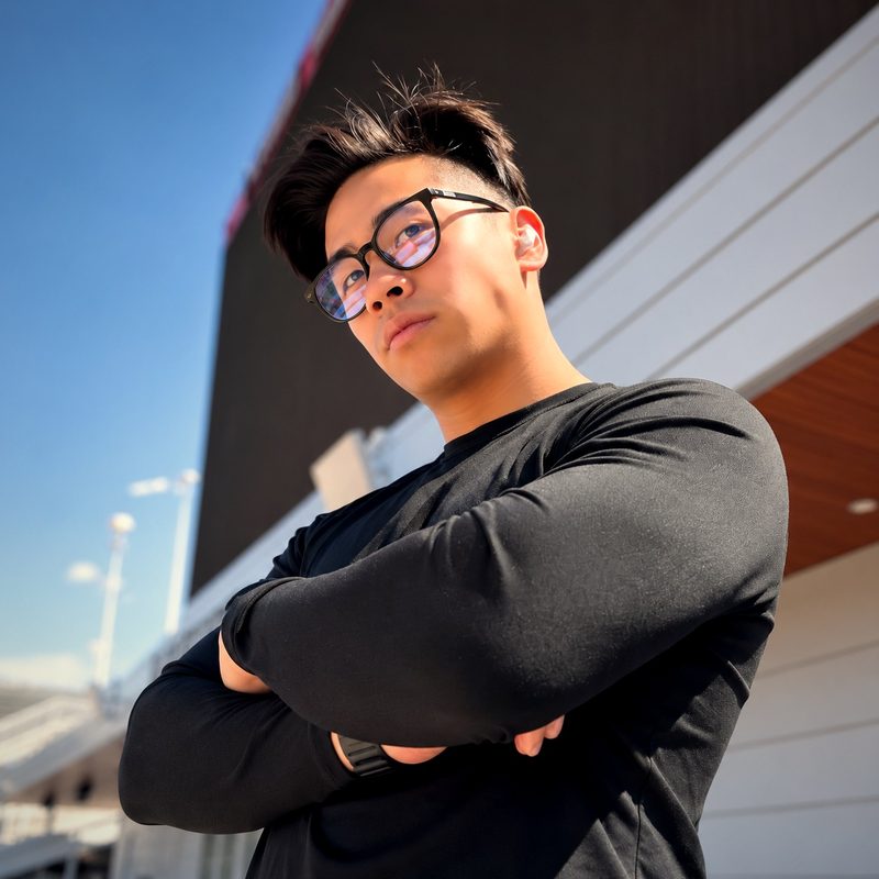 Evan Kwan in glasses and a black long-sleeve shirt, arms crossed, looking off to the side outside a stadium on a sunny day